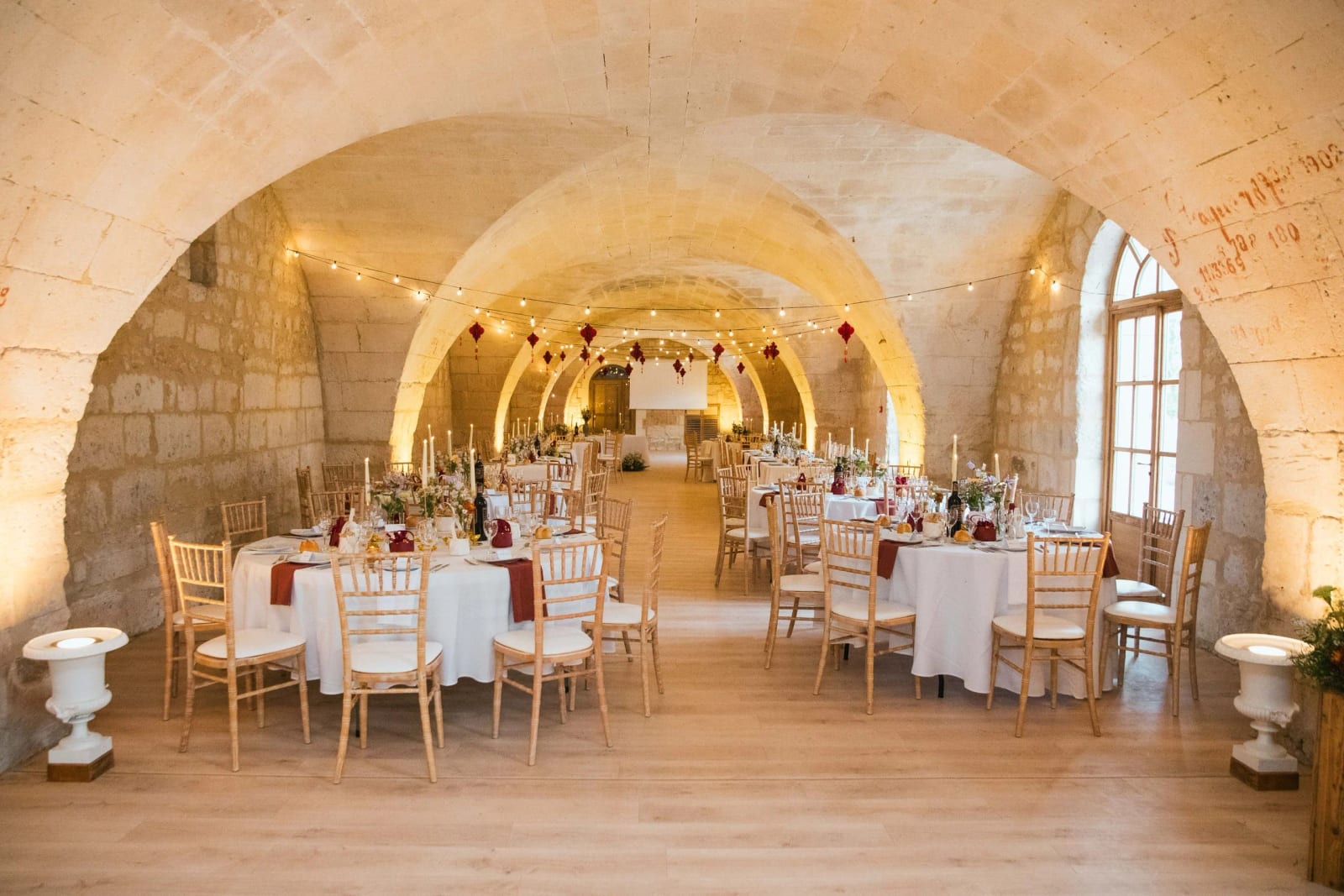 Salle voûtée en pierre du Domaine de Brion dressée de tables rondes, guirlandes lumineuses et lanternes rouges suspendues, mariage au Domaine de Brion, par Jean-Baptiste Argyre (Jaysphotography)