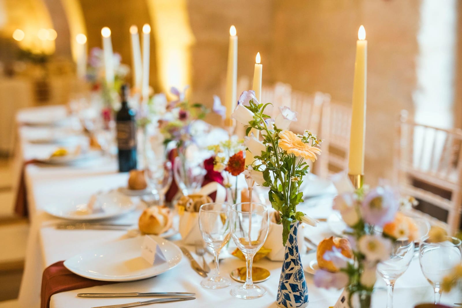 Table dressée sous les voûtes : hautes bougies, petits vases de fleurs, vase bleu et blanc et serviettes couleur rouille, mariage au Domaine de Brion, par Jean-Baptiste Argyre (Jaysphotography)