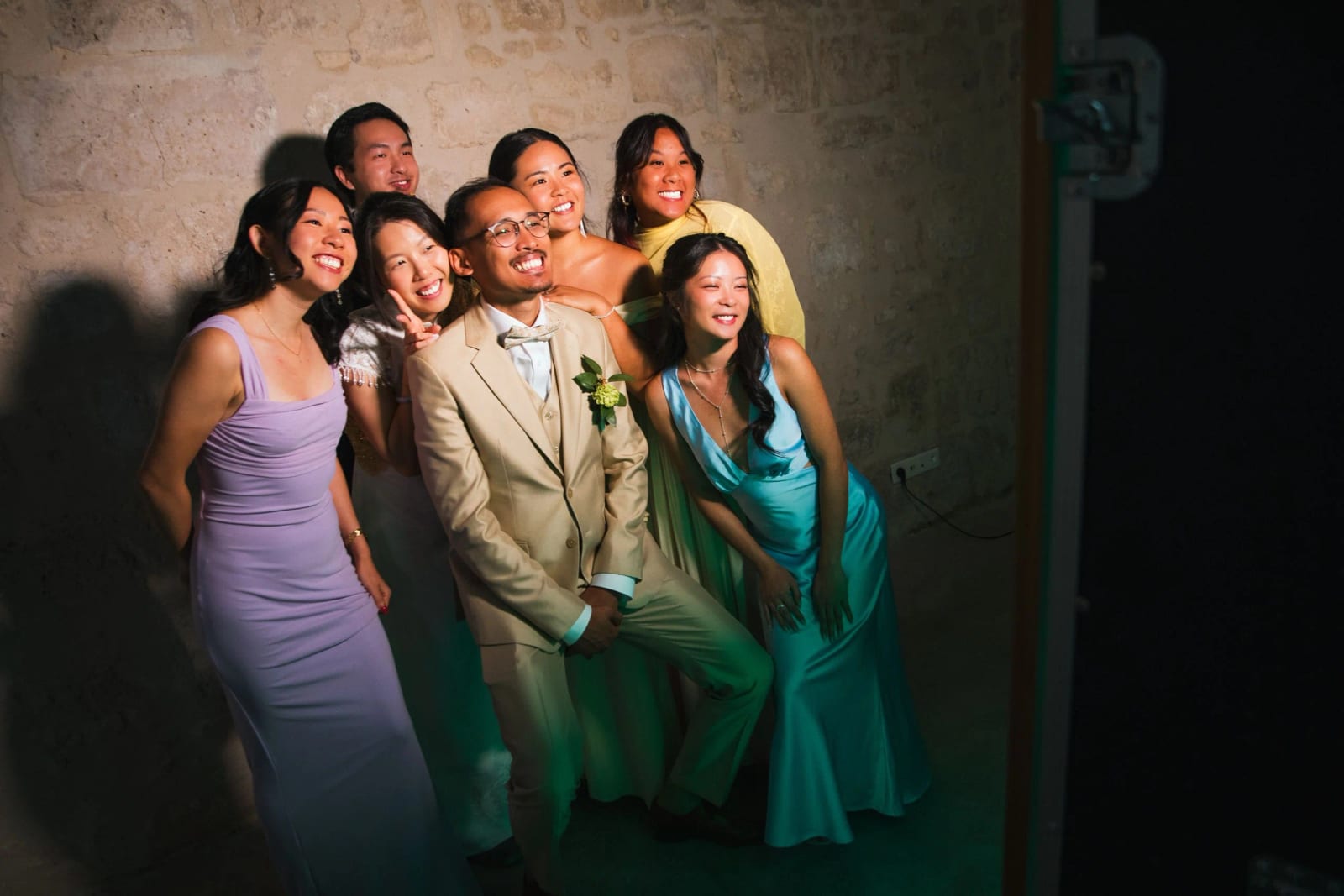 Le marié accroupi au milieu de ses amis en robes de couleur, éclairés par un projecteur contre un mur de pierre, mariage au Domaine de Brion, par Jean-Baptiste Argyre (Jaysphotography)