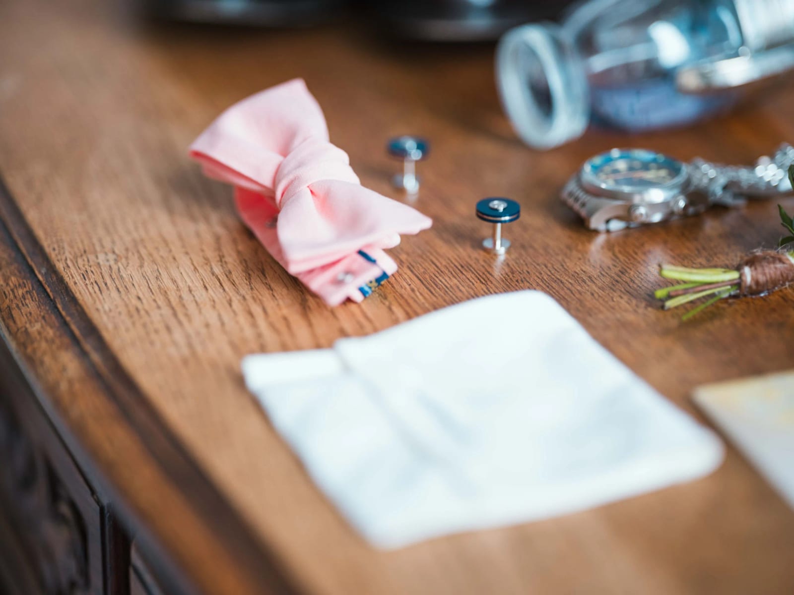 Accessoires du marié sur une commode de bois : nœud papillon rose poudré, boutons de manchette, montre, pochette et boutonnière, Château de Reilly, mariage au Château de Reilly, par Jean-Baptiste Argyre (Jaysphotography)