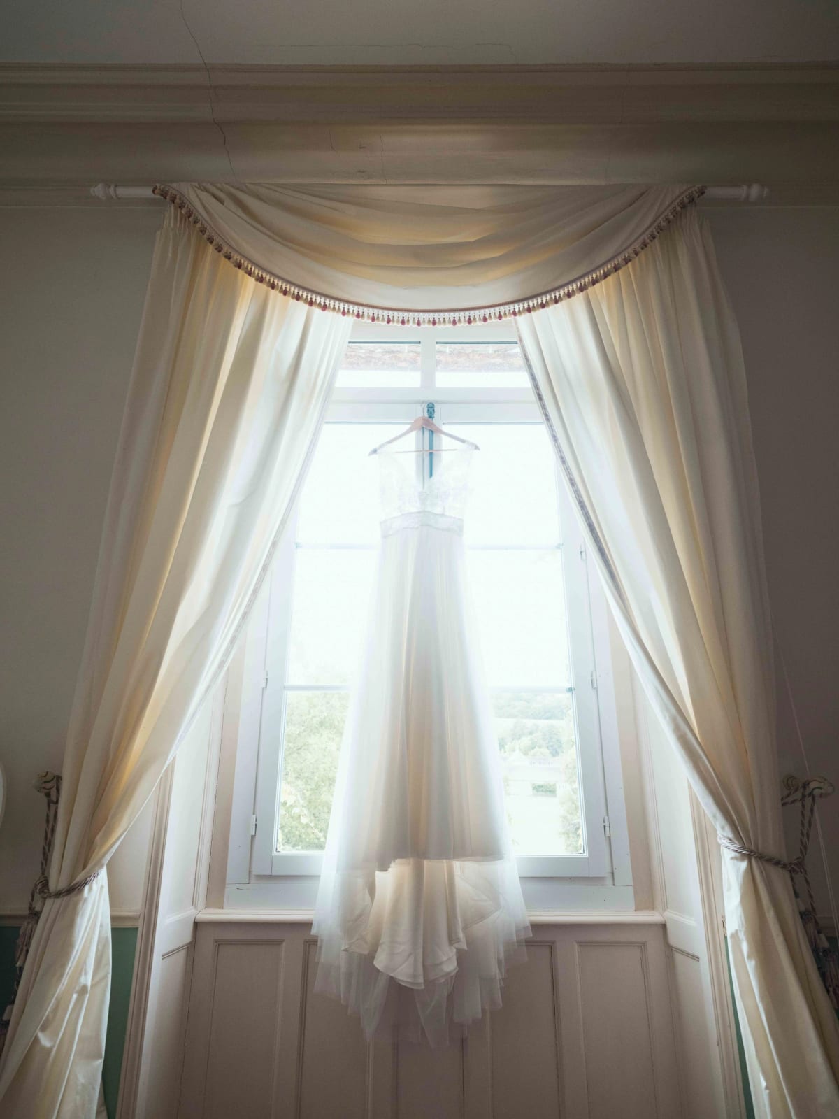 La robe de mariée suspendue devant une haute fenêtre drapée de rideaux crème, Château de Reilly, mariage au Château de Reilly, par Jean-Baptiste Argyre (Jaysphotography)