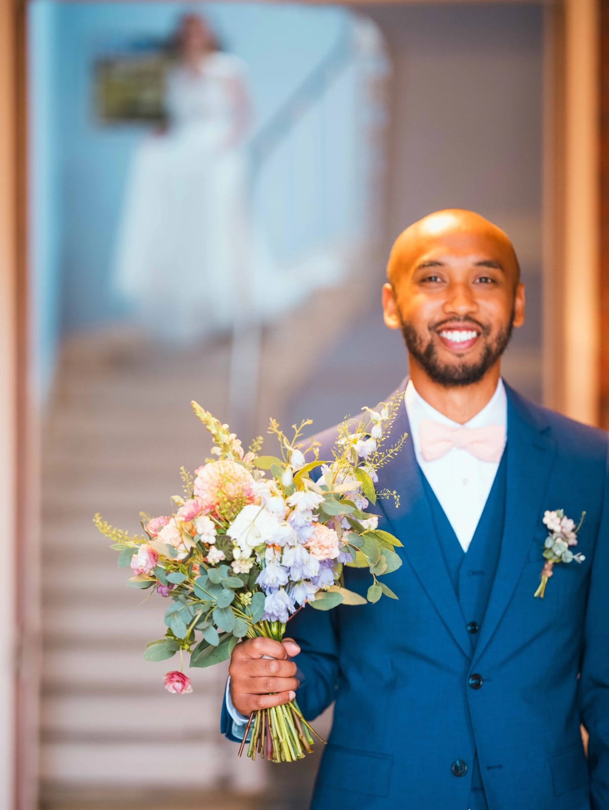 First look : le marié attend en bas de l'escalier avec le bouquet pastel, la mariée floue descend derrière lui, mariage au Château de Reilly, par Jean-Baptiste Argyre (Jaysphotography)