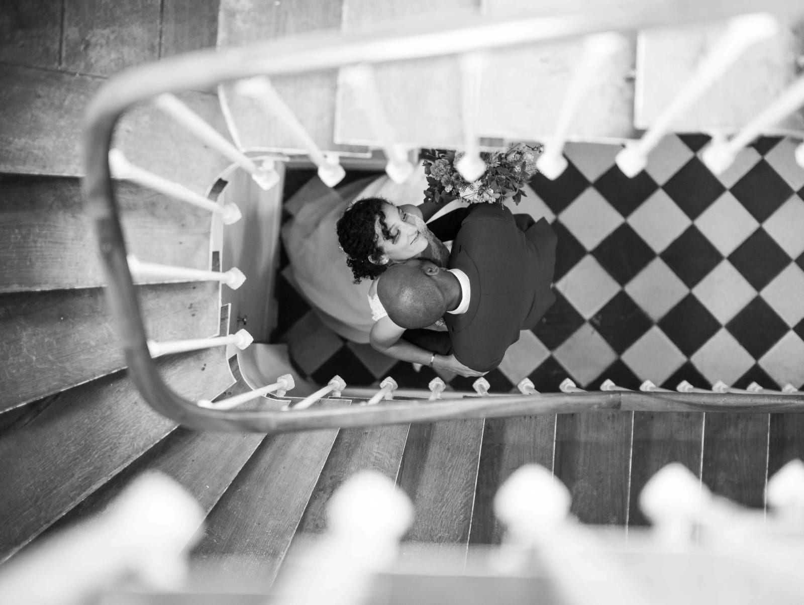 Vue plongeante en noir et blanc sur l'escalier en colimaçon à balustres blancs, les mariés s'embrassent sur le damier du sol, mariage au Château de Reilly, par Jean-Baptiste Argyre (Jaysphotography)