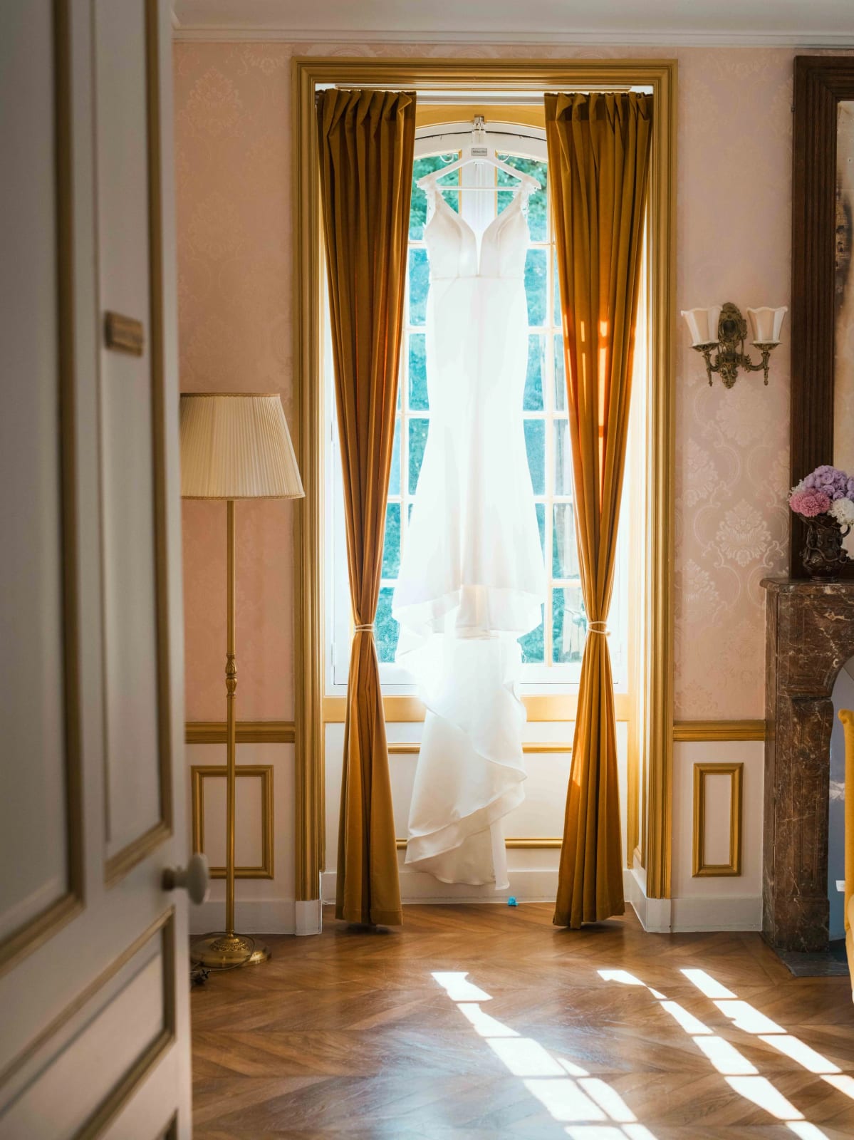 La robe de mariée suspendue à une haute fenêtre entre deux rideaux ocre, salon à parquet et moulures du Château de Moliens, mariage au Château de Moliens, par Jean-Baptiste Argyre (Jaysphotography)
