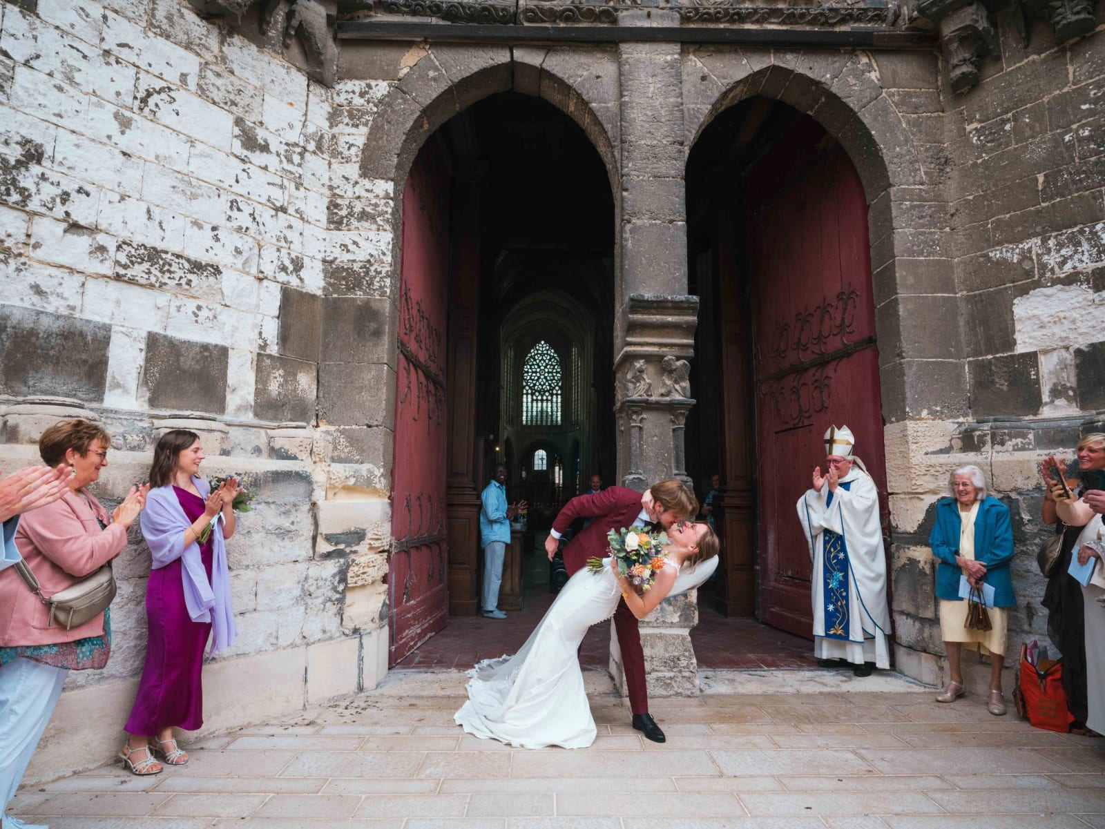 Sortie d'église sous le double portail de pierre de Saint-Étienne de Beauvais : le marié penche la mariée pour l'embrasser, l'évêque en mitre et chape à droite, invités qui applaudissent, mariage au Château de Moliens, par Jean-Baptiste Argyre (Jaysphotography)