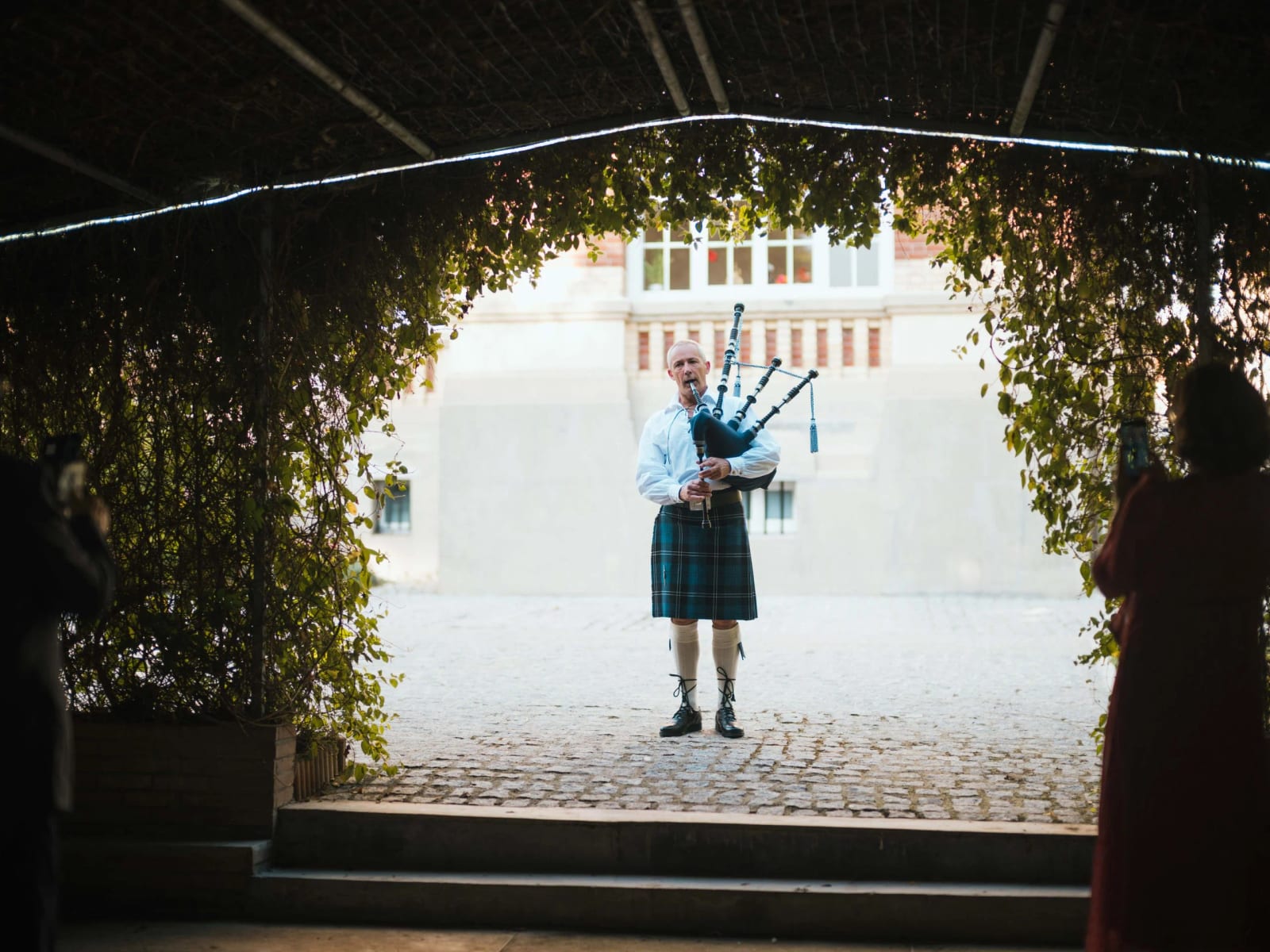 Un joueur de cornemuse en kilt seul dans la cour pavée du Château de Moliens, vu depuis la tonnelle de verdure, mariage au Château de Moliens, par Jean-Baptiste Argyre (Jaysphotography)