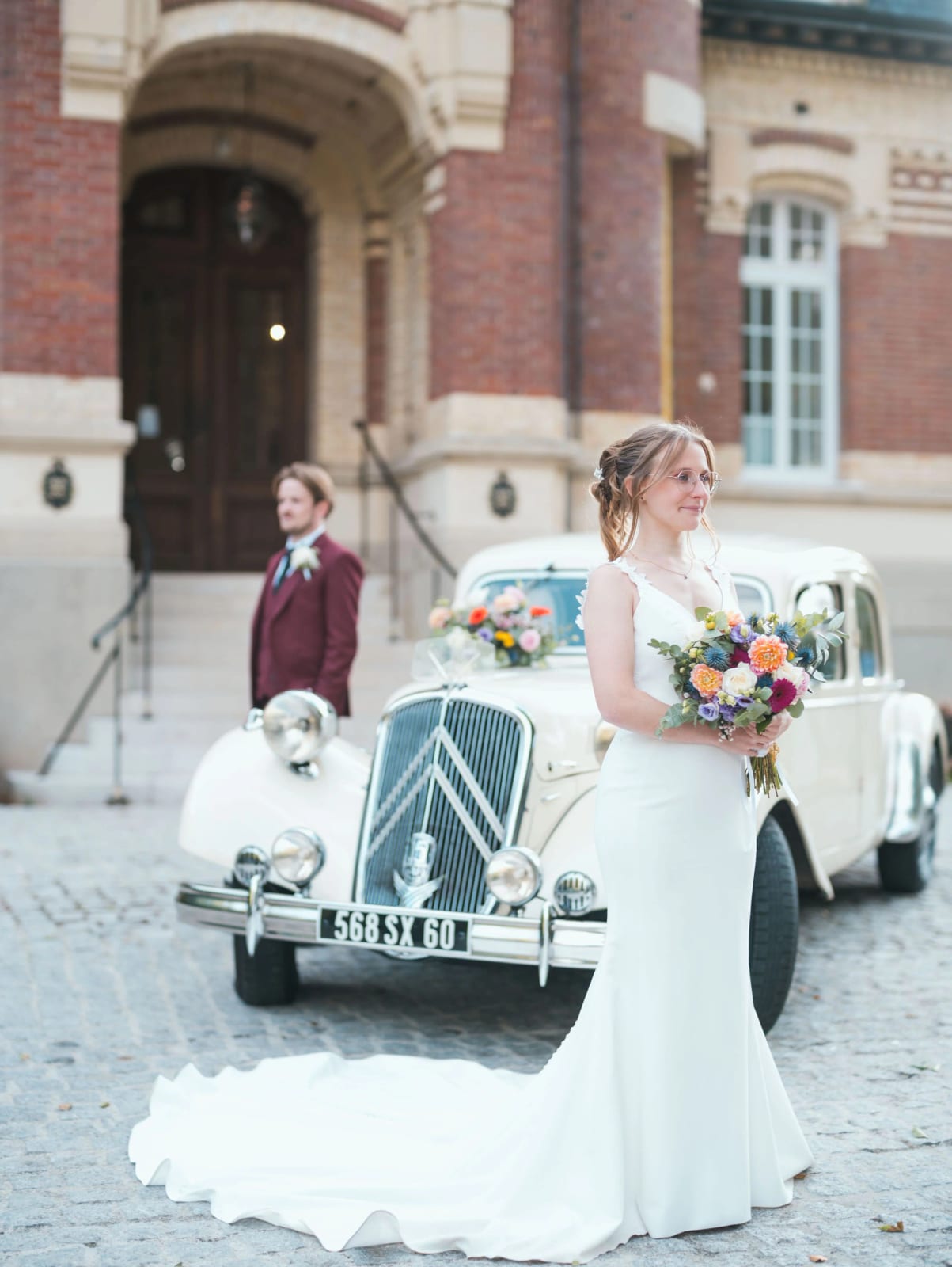 La mariée devant la Citroën Traction Avant blanche, traîne déployée sur les pavés, le marié sur le perron du Château de Moliens, mariage au Château de Moliens, par Jean-Baptiste Argyre (Jaysphotography)