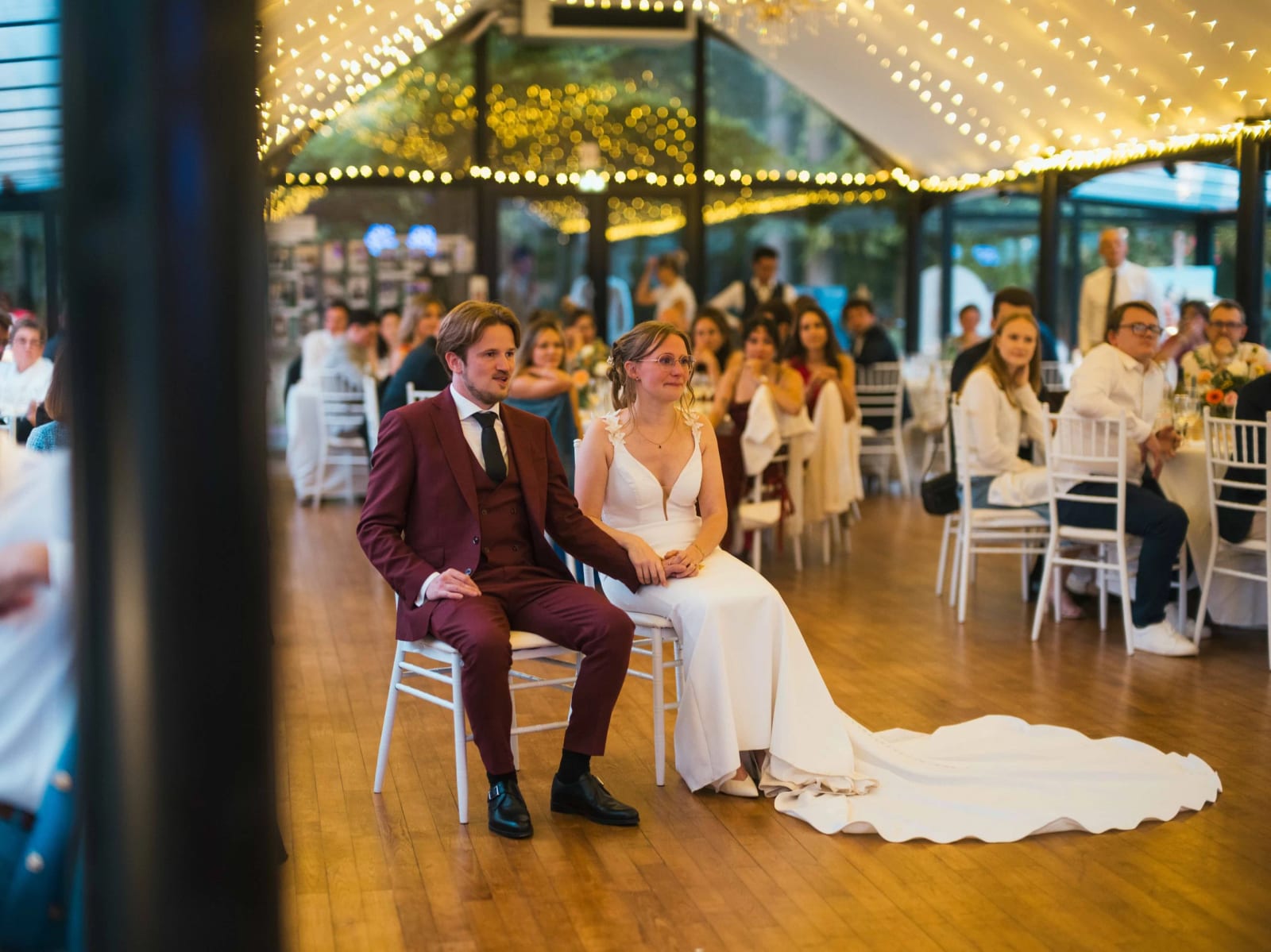 Les mariés assis sous la verrière du Château de Moliens pendant la soirée, guirlandes d'ampoules et tables des invités, mariage au Château de Moliens, par Jean-Baptiste Argyre (Jaysphotography)