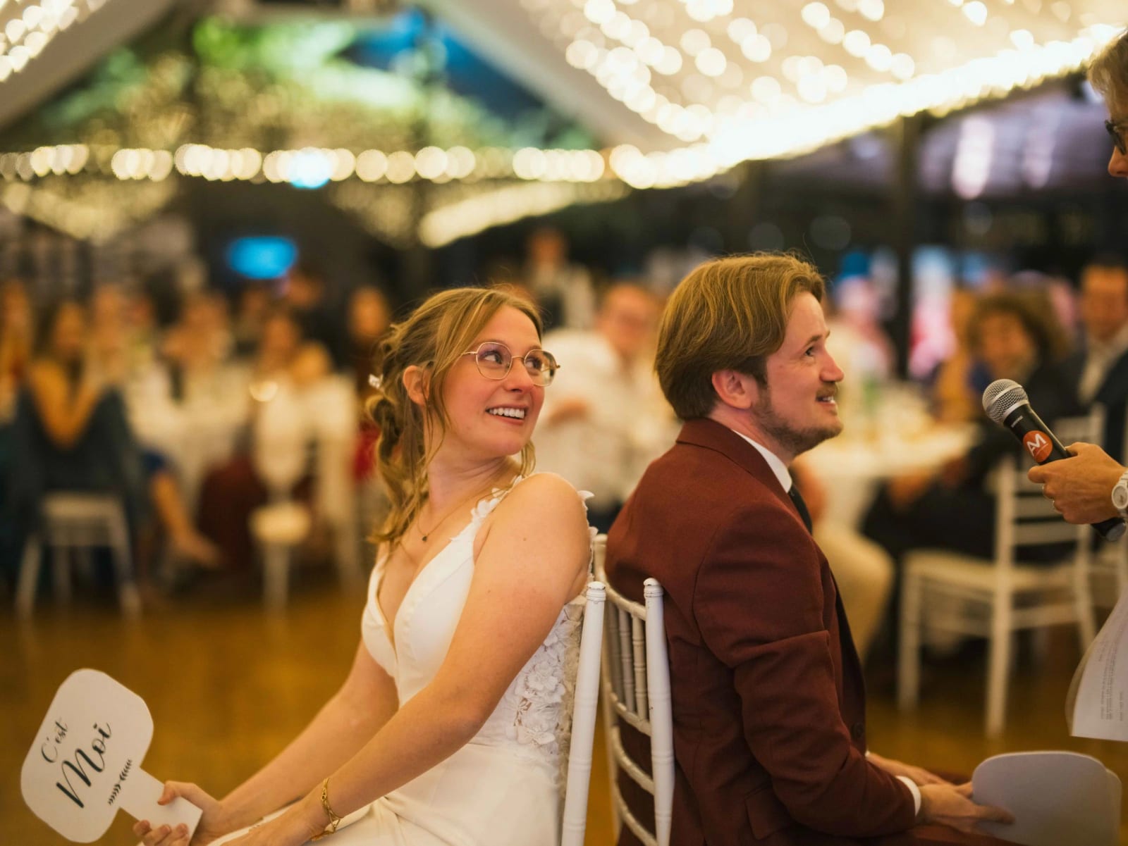 Le jeu des pancartes pendant le repas : la mariée rit en levant sa palette de bois, le marié détourne le regard, Château de Moliens, mariage au Château de Moliens, par Jean-Baptiste Argyre (Jaysphotography)