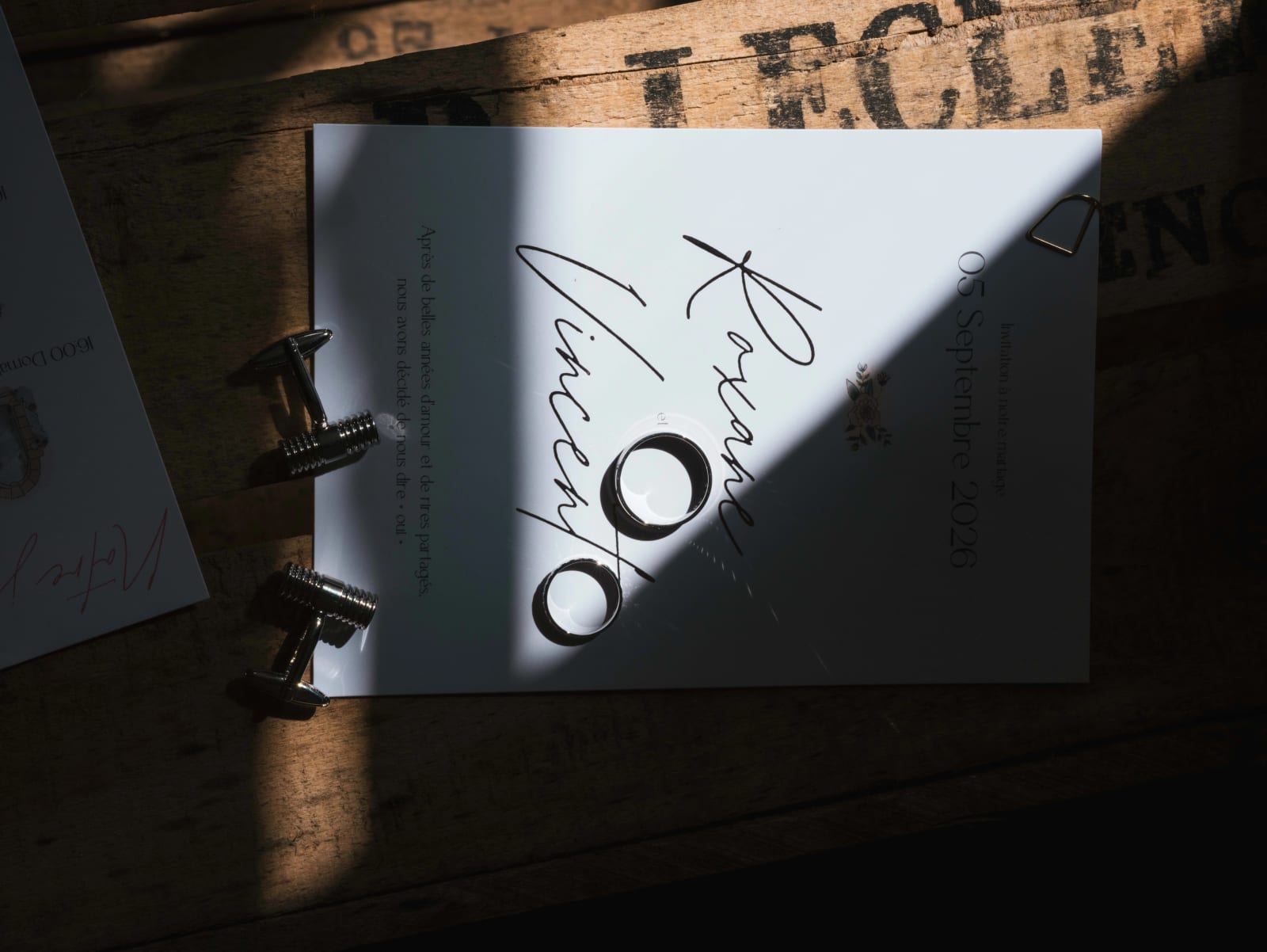 Faire-part « Roxane et Vincent, 05 Septembre 2026 » posé sur une caisse de bois, alliances et boutons de manchette dans un rai de soleil, mariage au Château de Sainte-Claire, Berneuil-sur-Aisne, par Jean-Baptiste Argyre (Jaysphotography)