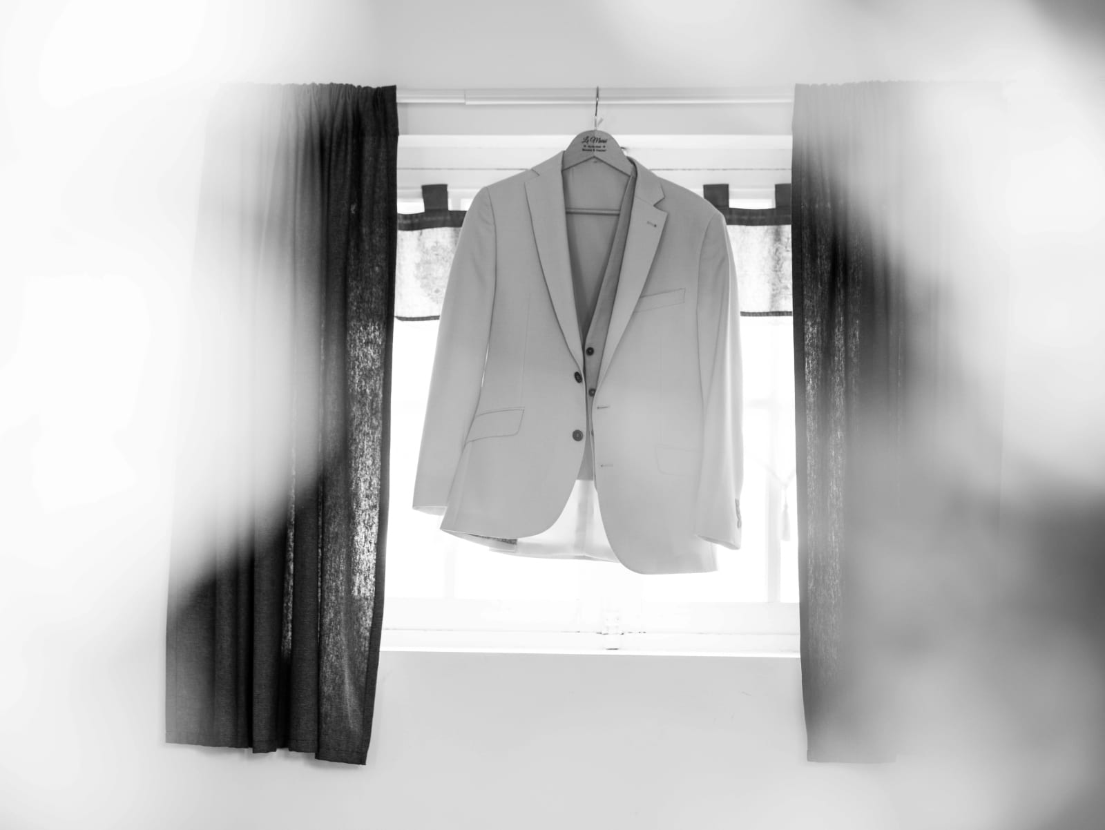 La veste du marié suspendue devant une fenêtre entre deux rideaux sombres, en noir et blanc, mariage au Château de Sainte-Claire, Berneuil-sur-Aisne, par Jean-Baptiste Argyre (Jaysphotography)