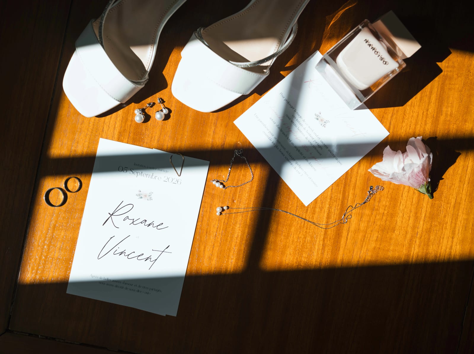 Accessoires de la mariée sur une table de bois : sandales blanches à strass, boucles d'oreilles en perles, alliances, faire-part, parfum et fleur rose, mariage au Château de Sainte-Claire, Berneuil-sur-Aisne, par Jean-Baptiste Argyre (Jaysphotography)