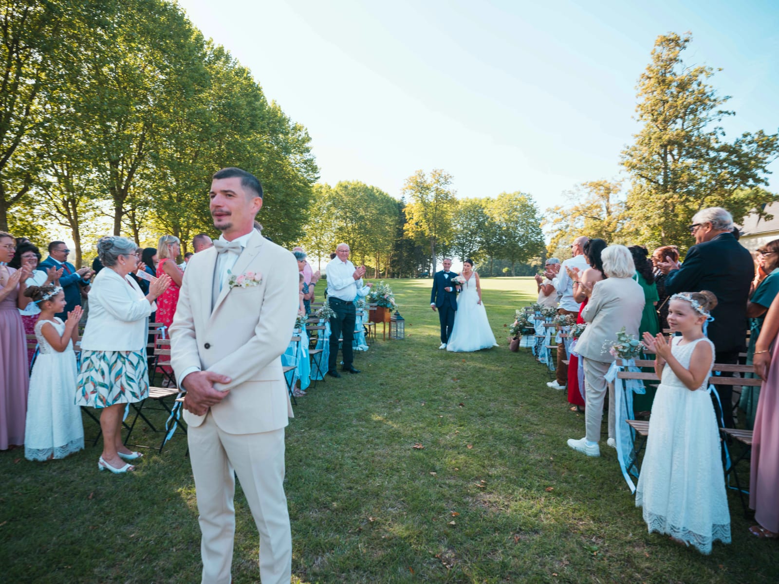 Entrée de la mariée au bras d'un proche dans l'allée de la cérémonie laïque, le marié de dos au premier plan, invités qui applaudissent, mariage au Château de Sainte-Claire, Berneuil-sur-Aisne, par Jean-Baptiste Argyre (Jaysphotography)