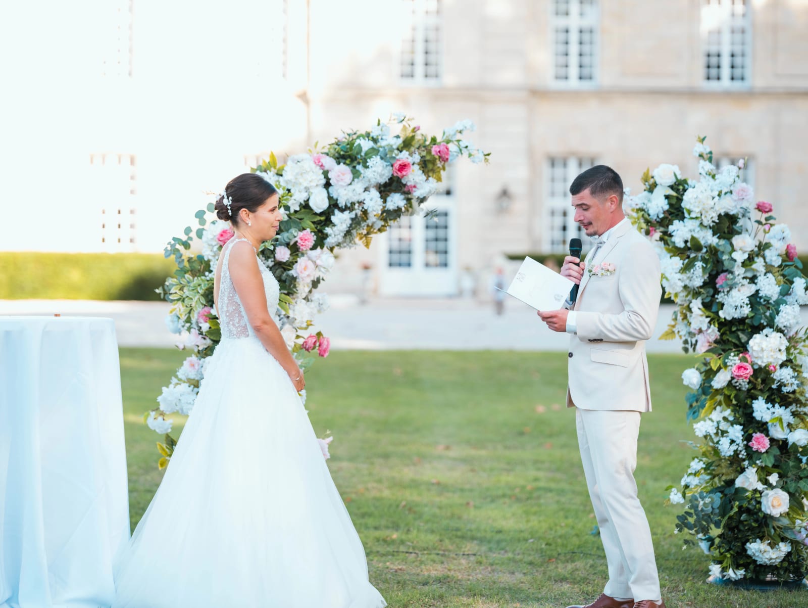 Le marié lit ses vœux au micro devant une arche de fleurs blanches et roses, façade du Château de Sainte-Claire en fond, mariage au Château de Sainte-Claire, Berneuil-sur-Aisne, par Jean-Baptiste Argyre (Jaysphotography)