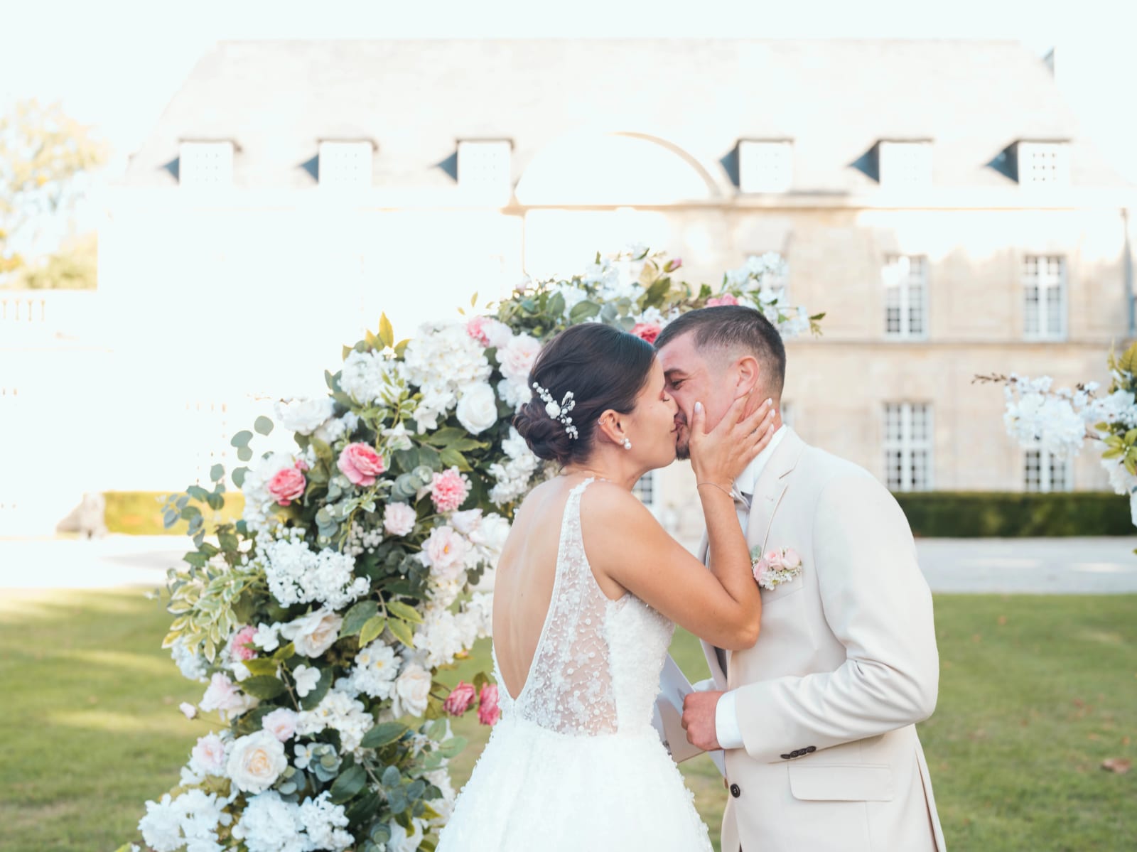 Baiser des mariés devant l'arche fleurie, dos ouvert de la robe en dentelle, château en arrière-plan, mariage au Château de Sainte-Claire, Berneuil-sur-Aisne, par Jean-Baptiste Argyre (Jaysphotography)