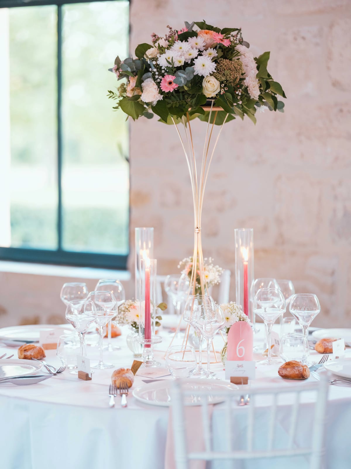 Table ronde avec centre de table sur pied doré, gerberas et marguerites, numéro de table rose et chaises blanches, mur de pierre, mariage au Château de Sainte-Claire, Berneuil-sur-Aisne, par Jean-Baptiste Argyre (Jaysphotography)