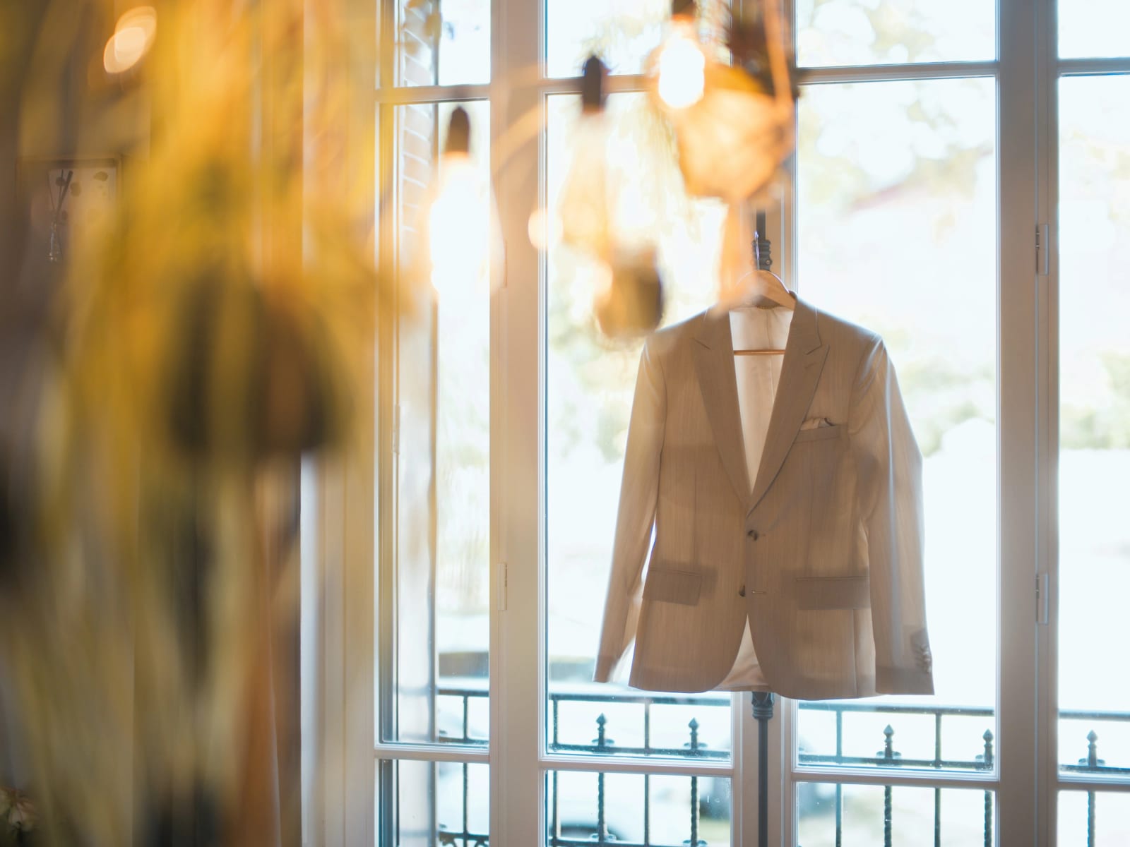 Veste de costume beige sur un cintre devant une porte-fenêtre à garde-corps en fer forgé, ampoules suspendues au premier plan, préparatifs du marié, mariage au Clos d'Amblaincourt, par Jean-Baptiste Argyre (Jaysphotography)