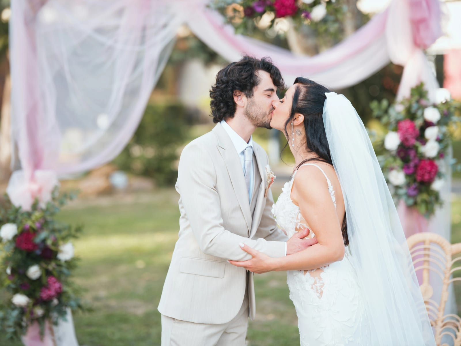 Baiser des mariés sous l'arche de cérémonie drapée de tulle rose et garnie de roses, mariage au Clos d'Amblaincourt, par Jean-Baptiste Argyre (Jaysphotography)