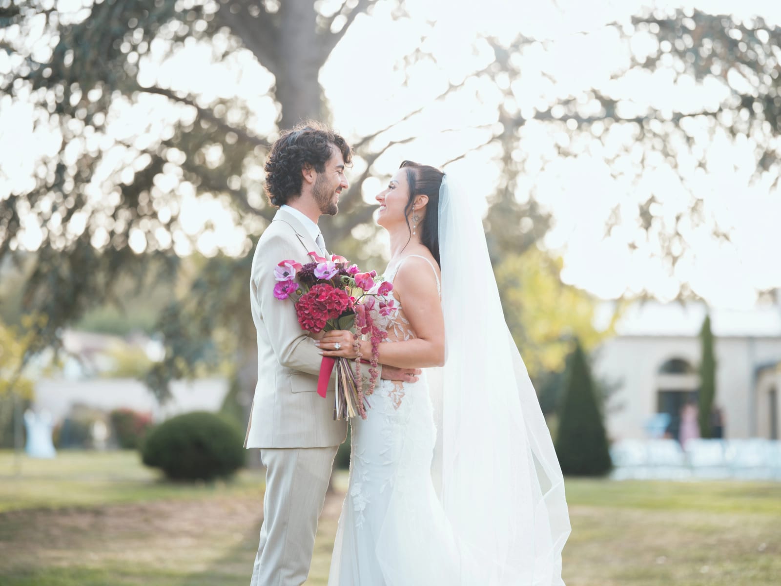Les mariés face à face sous un grand cèdre, bouquet rouge et rose, chaises blanches et bâtiment à fenêtres en arc au fond, mariage au Clos d'Amblaincourt, par Jean-Baptiste Argyre (Jaysphotography)