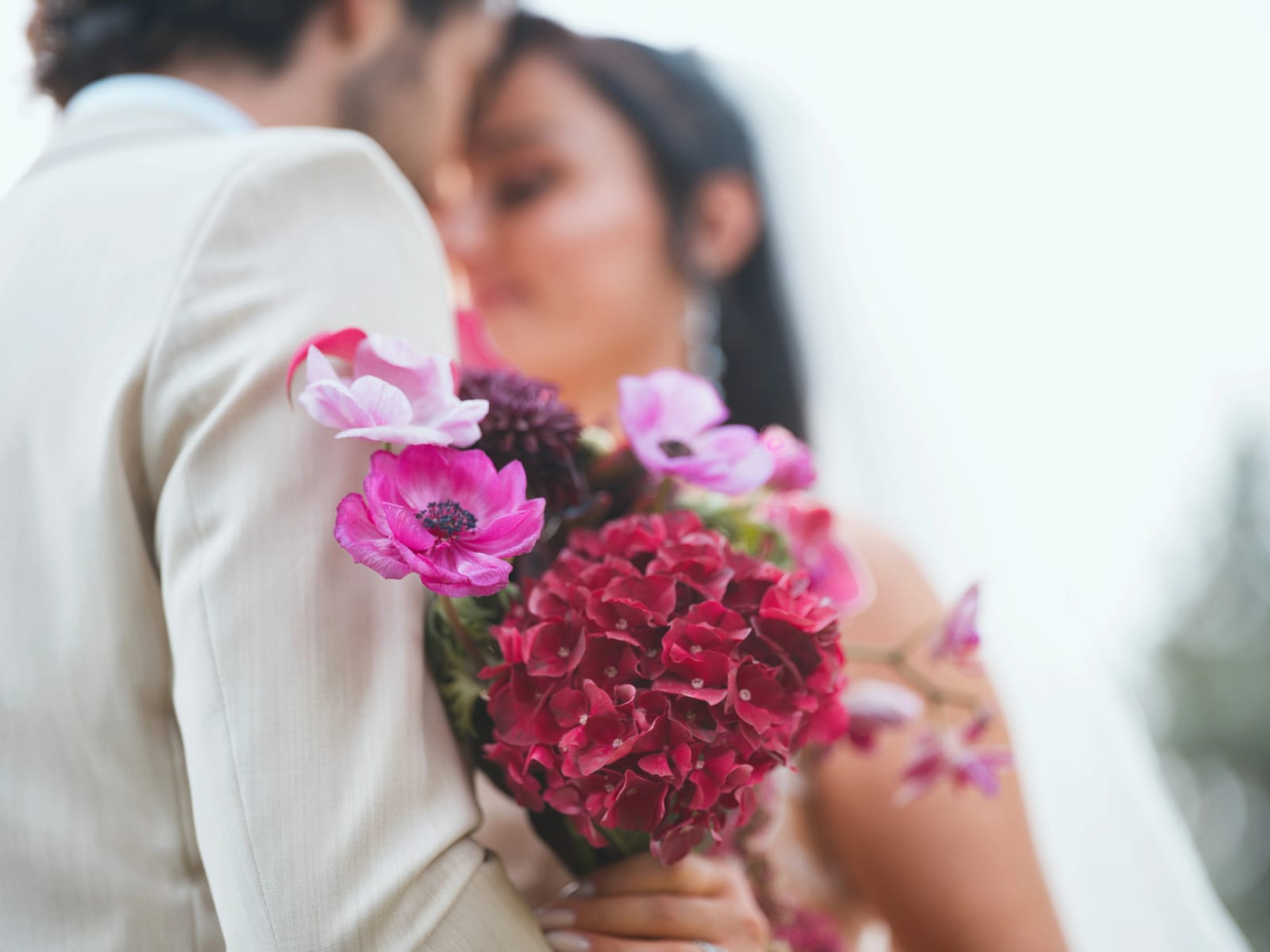 Bouquet d'anémones roses et d'hortensia rouge contre l'épaule du marié, mariés flous au second plan, mariage au Clos d'Amblaincourt, par Jean-Baptiste Argyre (Jaysphotography)