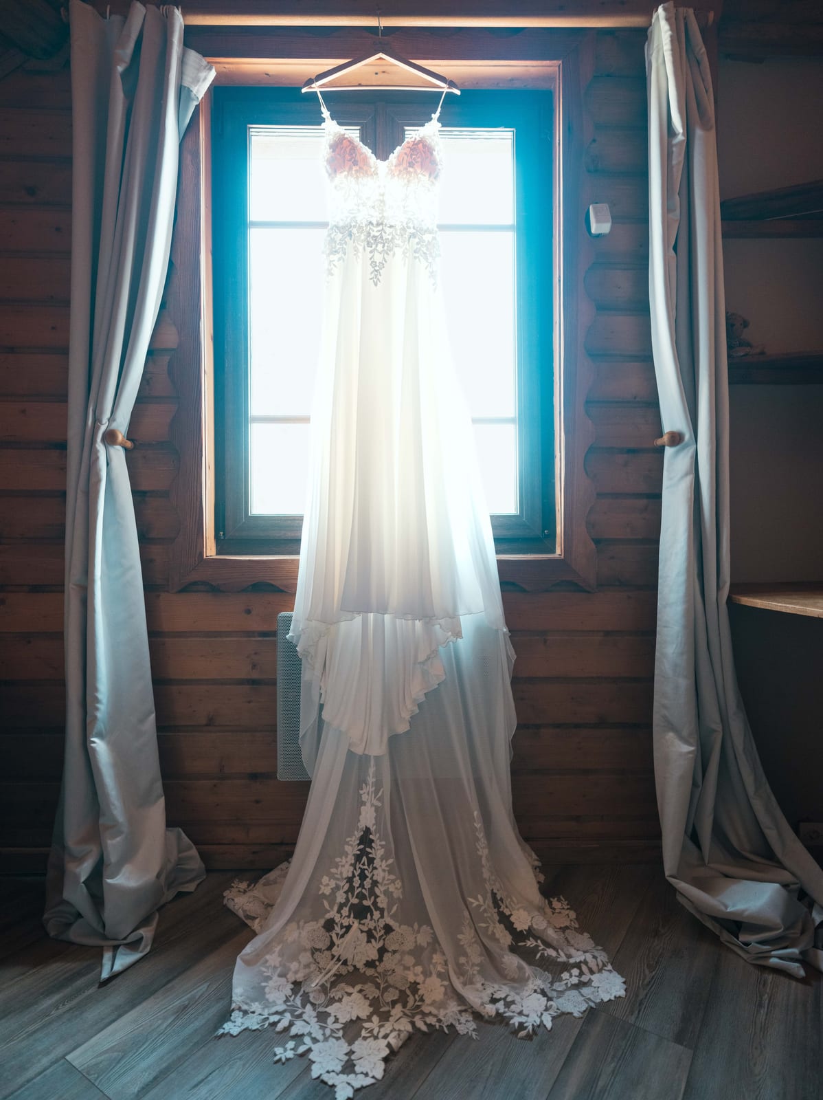 La robe de Mathilde accrochée devant la fenêtre à barreaux bleus du refuge, dentelle florale sur fond de lumière naturelle du matin, mariage au Refuges des 3 Ours, par Jean-Baptiste Argyre (Jaysphotography)