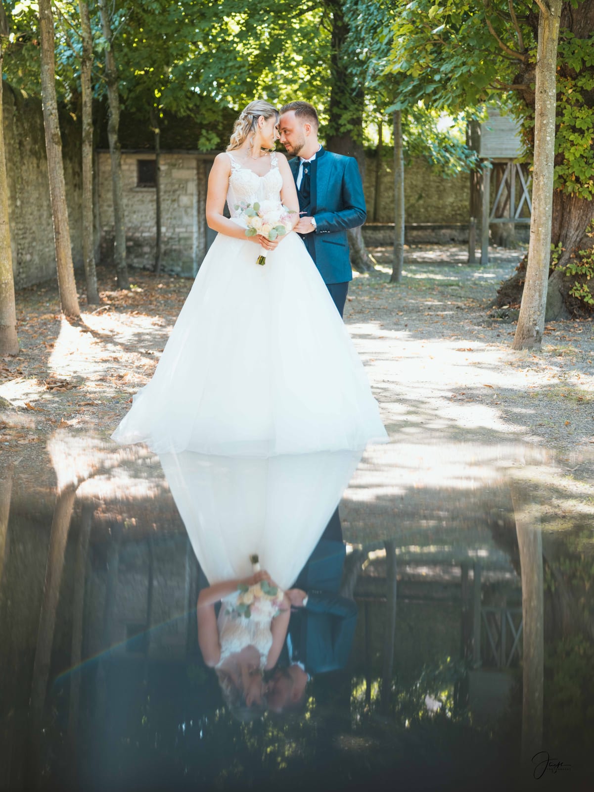 Ophélie et Vincent enlacés se reflétant dans un miroir en extérieur, robe blanche et costume bleu canard, parc en arrière-plan, mariage au Domaine de Monceaux, par Jean-Baptiste Argyre (Jaysphotography)