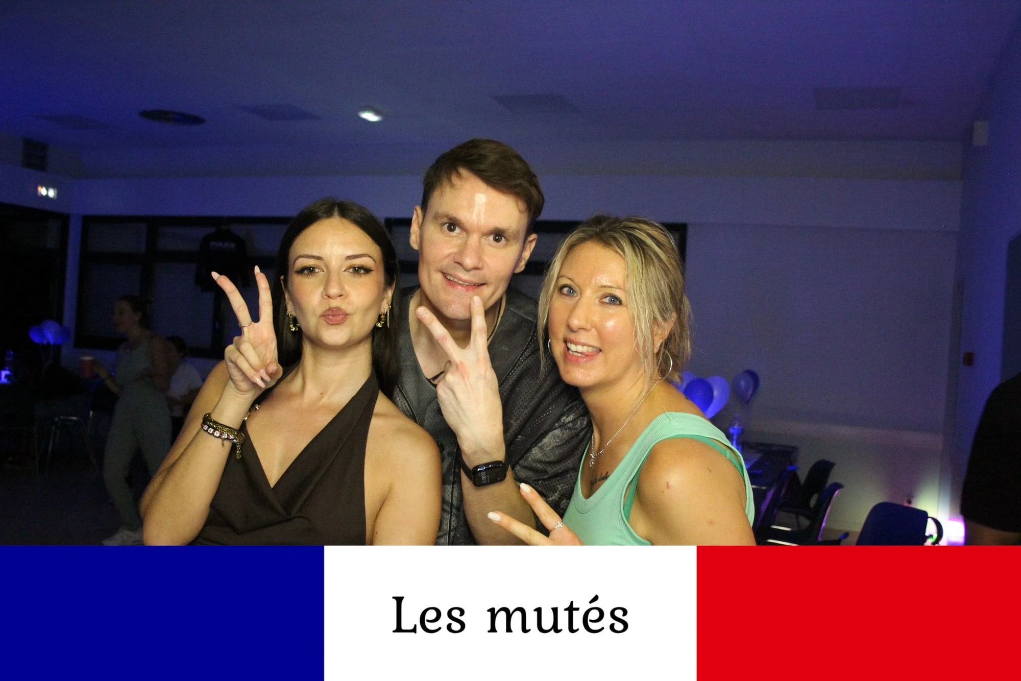 Deux invitées et un invité prennent la pose ensemble, doigts en V, ballons bleus et blancs au fond de la salle, mariage à la Salle des fêtes de Saint-Sauveur (Oise), par Jean-Baptiste Argyre (Jaysphotography)