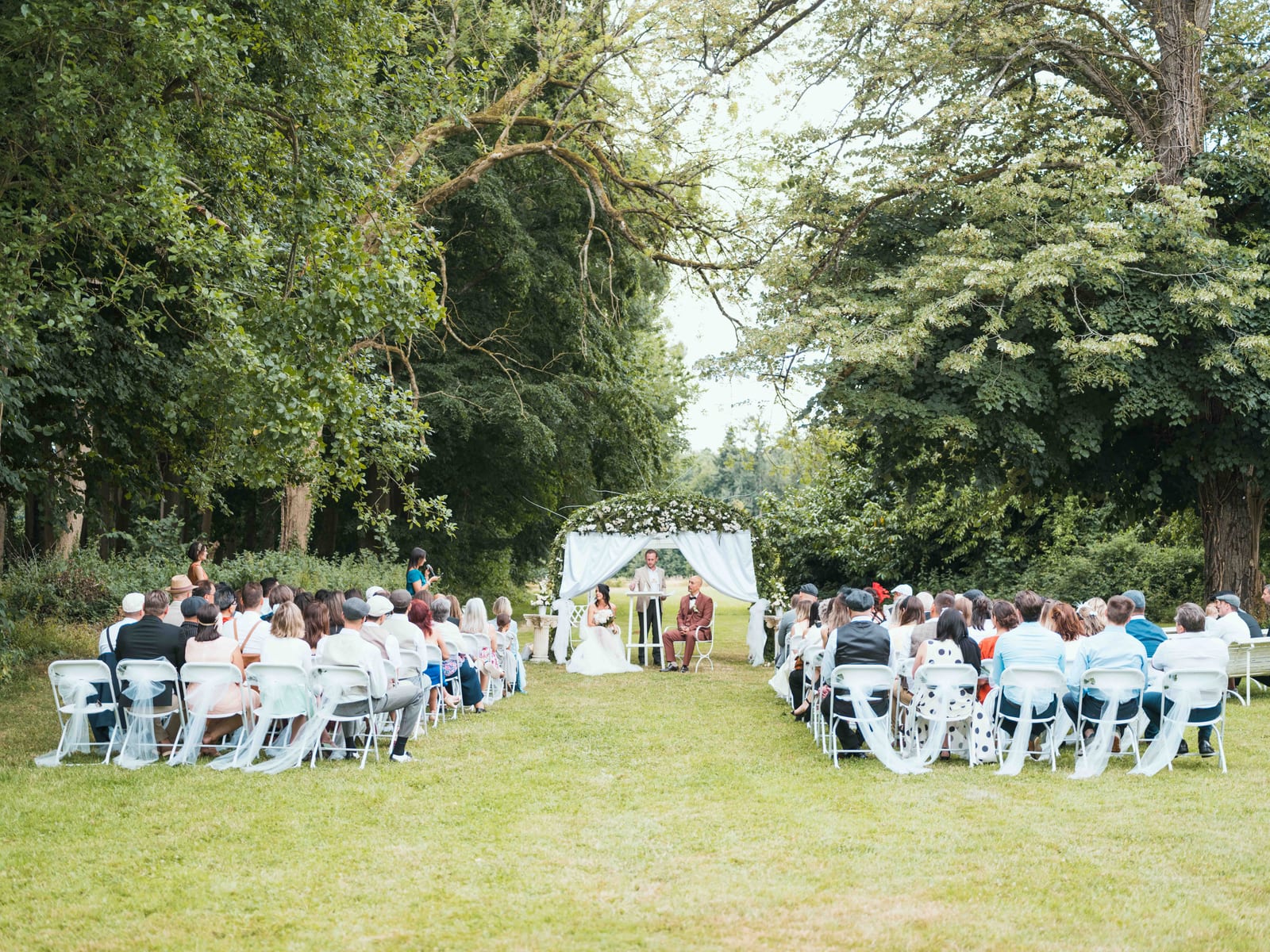 Vue d'ensemble de la cérémonie laïque dans les jardins du château, invités assis face à l'arche fleurie, mariage au Château de la Houssoye, par Jean-Baptiste Argyre (Jaysphotography)