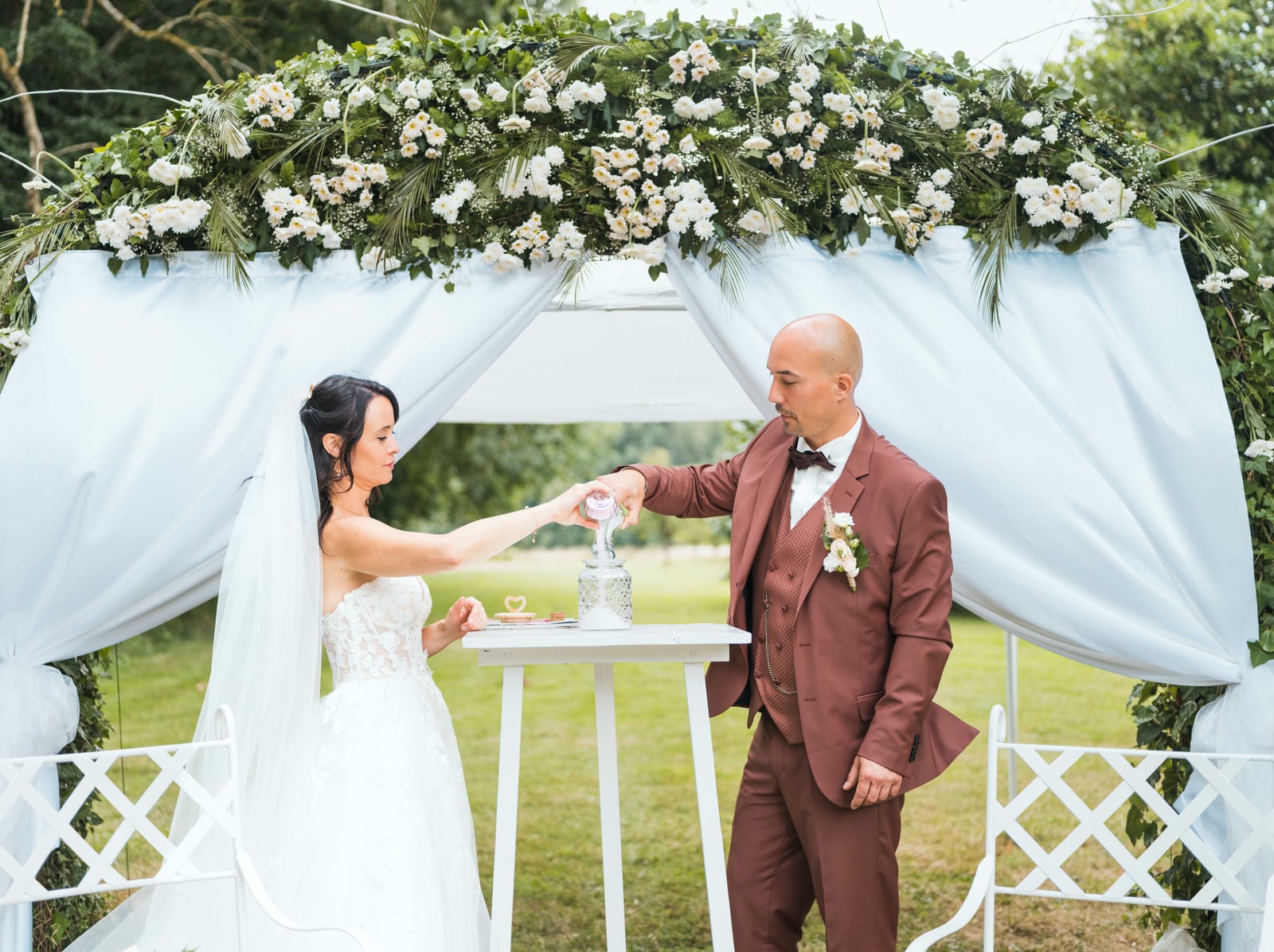 Stéphanie et Fabien devant l'arche blanche fleurie, échange de regards pendant la cérémonie, mariage au Château de la Houssoye, par Jean-Baptiste Argyre (Jaysphotography)