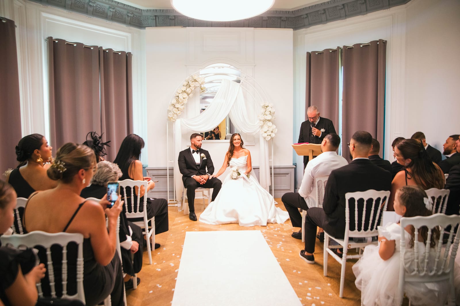 Wendy et Daniel devant le portail en arche du château, robe blanche et smoking noir en contraste sur la pierre néoclassique, mariage au Château de Lamorlaye, par Jean-Baptiste Argyre (Jaysphotography)